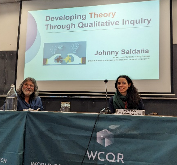 A man and a woman sitting along a table, with a screen in background, that reads - Developing Theory Through Qualitative Inquiry, during the final plenary of the WCQR. 