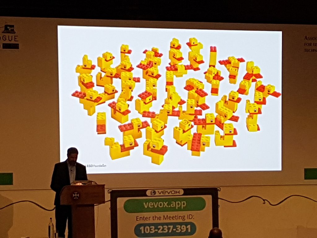 A man on a stage with a presentation slide showing several Lego ducks built in different configurations of bricks. 