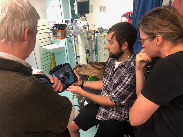 Three people standing in a hospital bed bay, looking at an ipad screen.