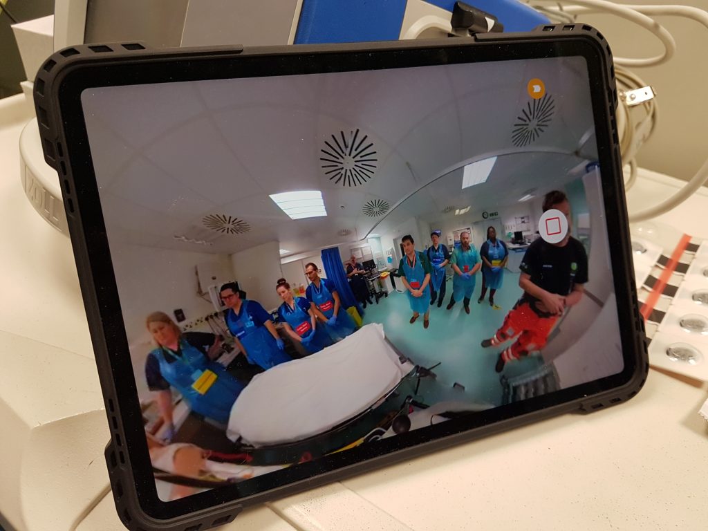 An ipad resting on a hospital trolley. The screen shows a fish eye view of trauma staff gathered around a bed. 