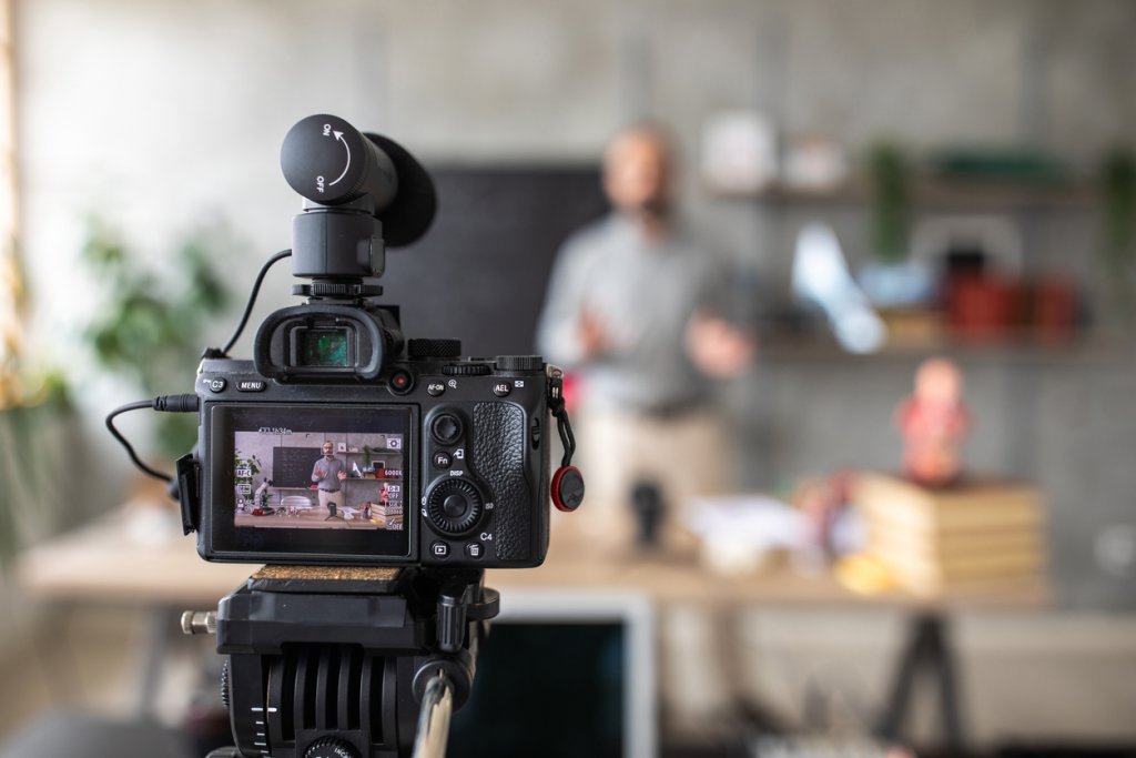 DSLR camera on tripod, recording a video of teacher. He is in the background of the shot, standing behind a table, out of focus.
