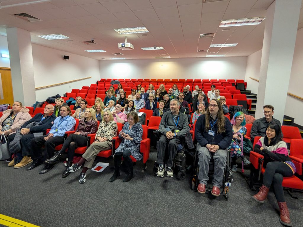 Group photo of approximately 40 presenters and organisers seated in a lecture theatre with red tiered seating. Attendees include wheelchair users in the front row.