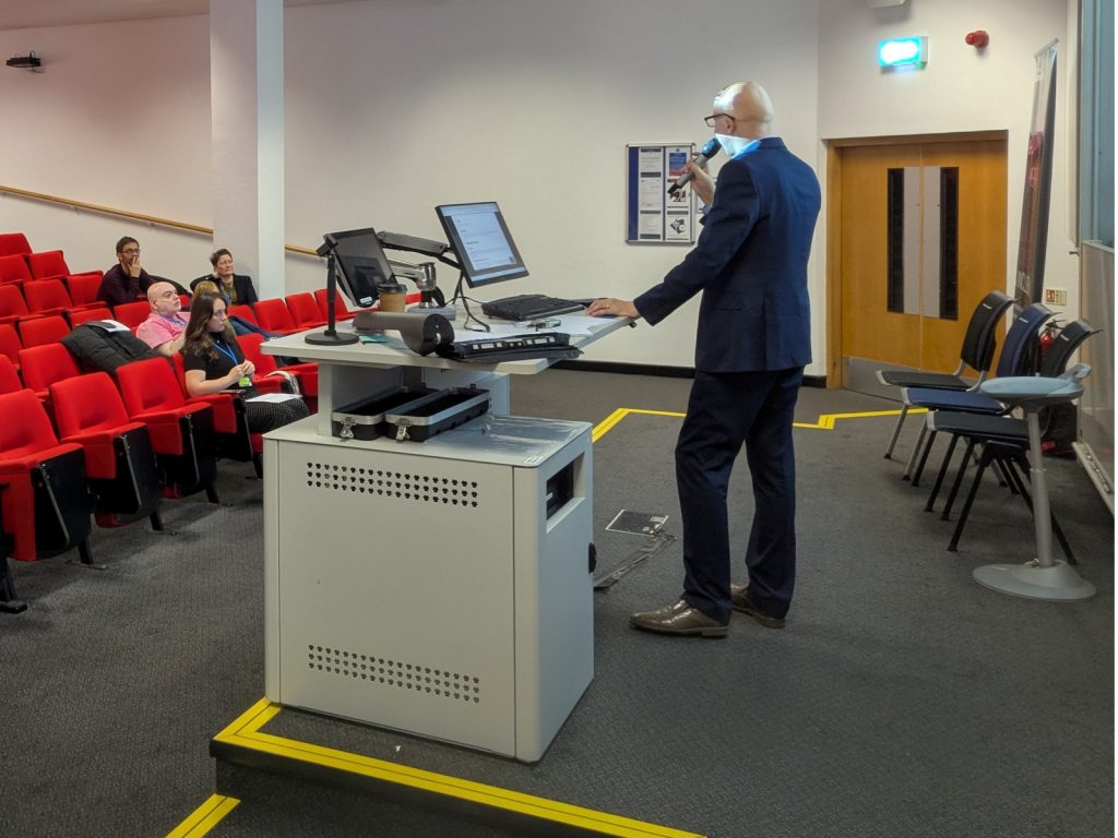 Matthew Deeprose standing at a presentation lectern with dual monitors, holding a microphone and presenting to attendees seated in red tiered seating.