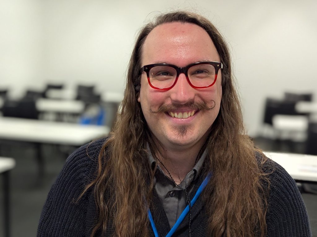 Piers Wilkinson smiling at the camera in a seminar room, wearing red-framed glasses, a dark cardigan, and a blue lanyard.