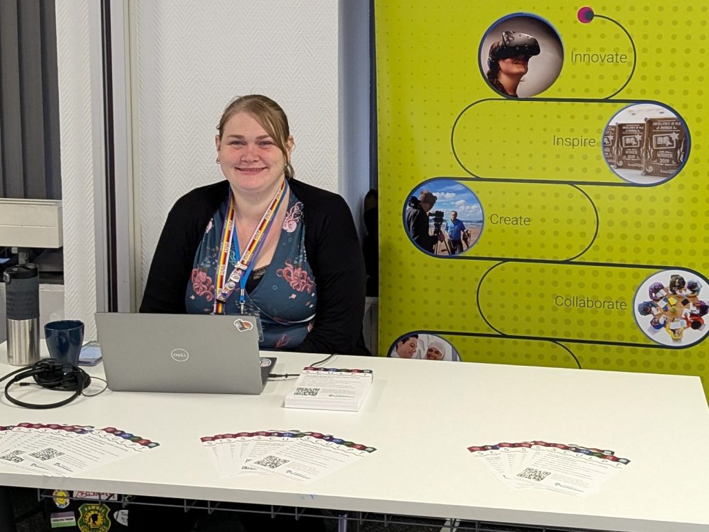 Stephanie Wright sitting at a table with information sheets laid out in front, a laptop, and a yellow display banner behind showing "Innovate, Inspire, Create, Collaborate".