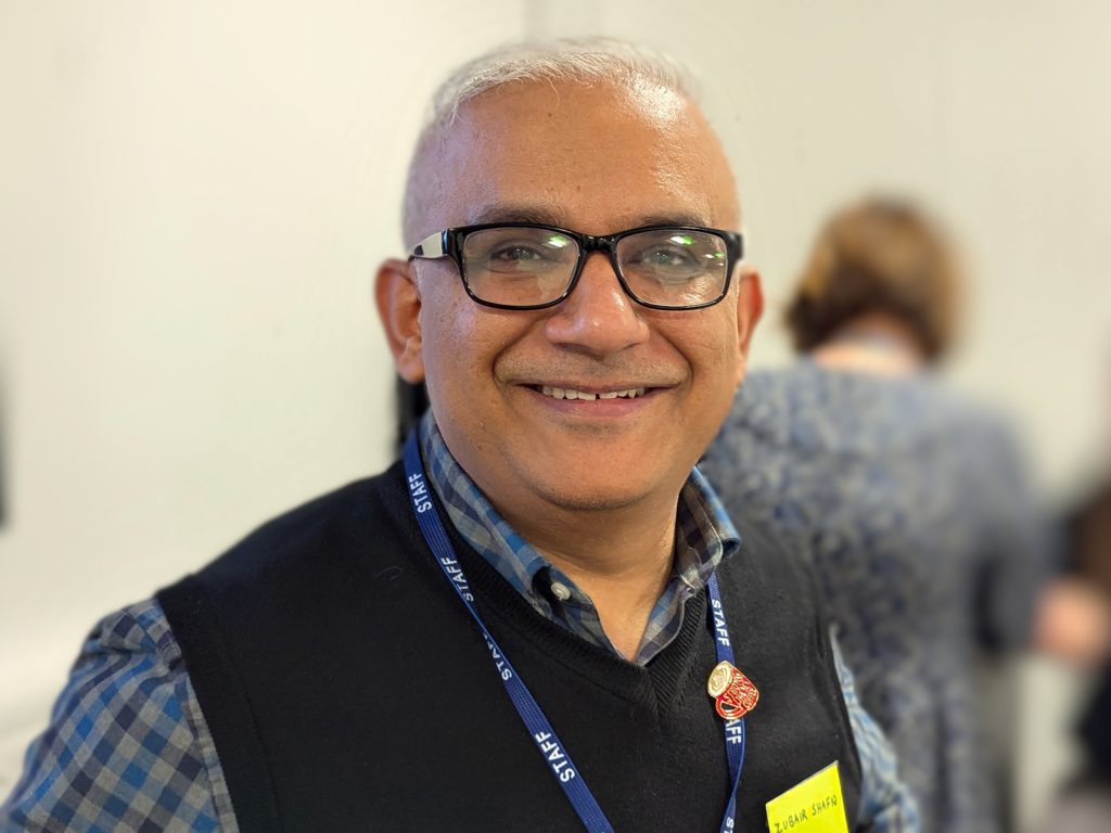 Dr Zubair Shafiq smiling at the camera, wearing black-framed glasses, a black sleeveless jumper over a checked shirt, and a blue staff lanyard.