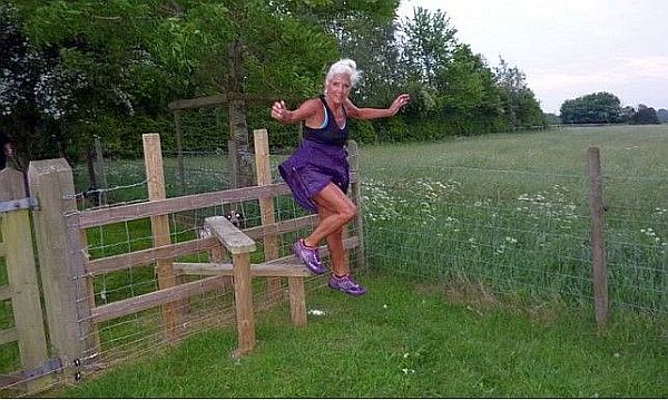 A silver haired woman wearing purple running atire is flamboyantly stepping off a rural style amidst grassy fields and tree-lined hedgerows.