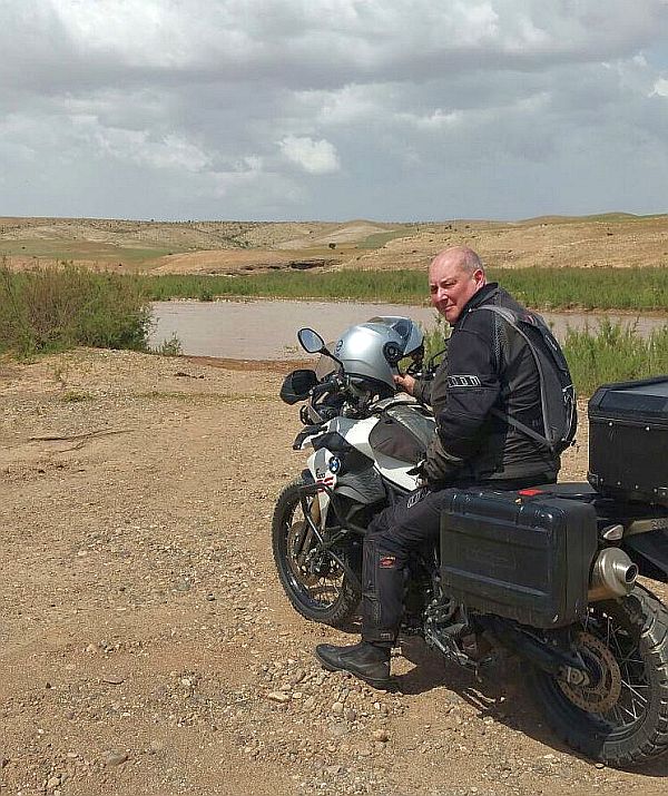A man astride a travelling motorcycle, leg parked overlooking a small lake and flat dunes towards an overcast horizon. The man is looking back at the photographer. 