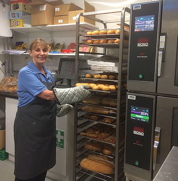 A proud women smiling towards the camera in a blue shirt, grey apron and  protective oven gloves, removing a tray of freshly based donuts from a cooking storage rack.