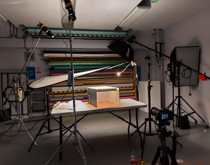 A professional photography studio setup featuring lighting equipment, a camera on a tripod, and a small object on a table being photographed against colorful backdrop rolls.