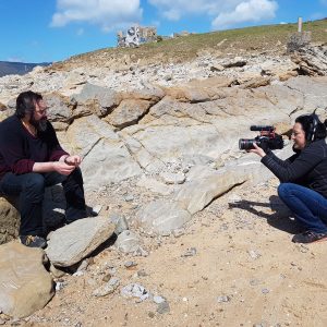 A man sitting on a rock whilst a woman points a camera at him. 