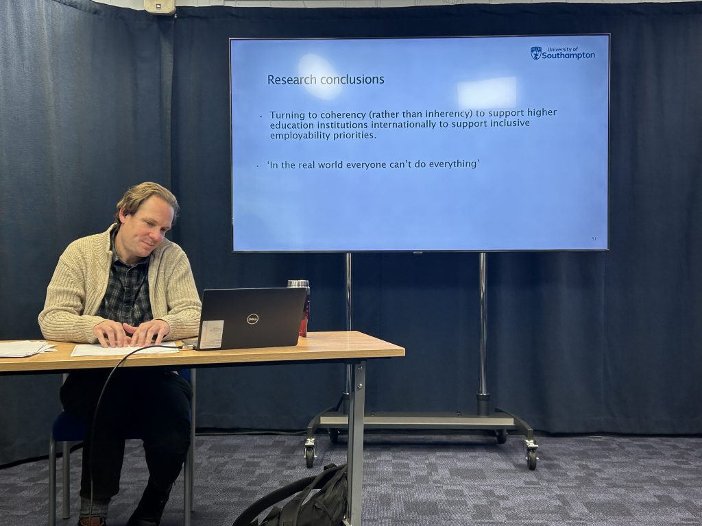 Dr Ben Whitburn sitting at a desk with a laptop and a projector screen, with information about his seminar. 