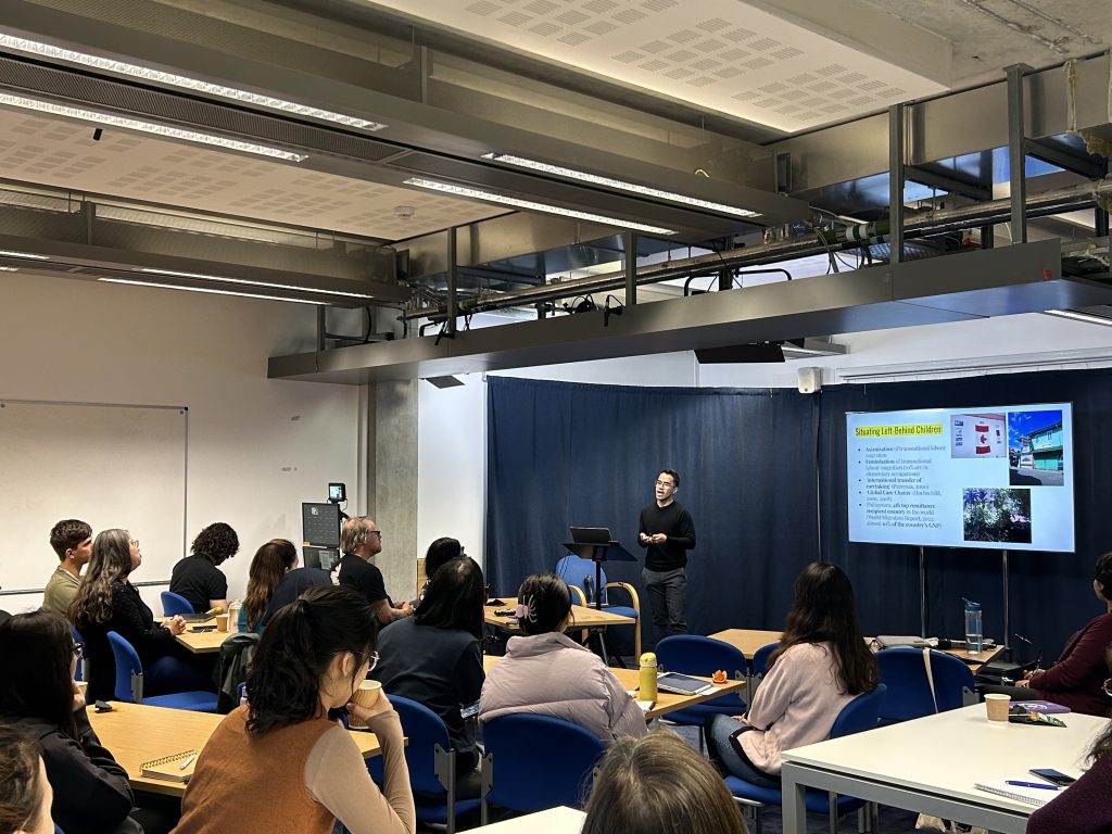 A person stood in front of a group of about 12 people presenting his slides with some images and text.