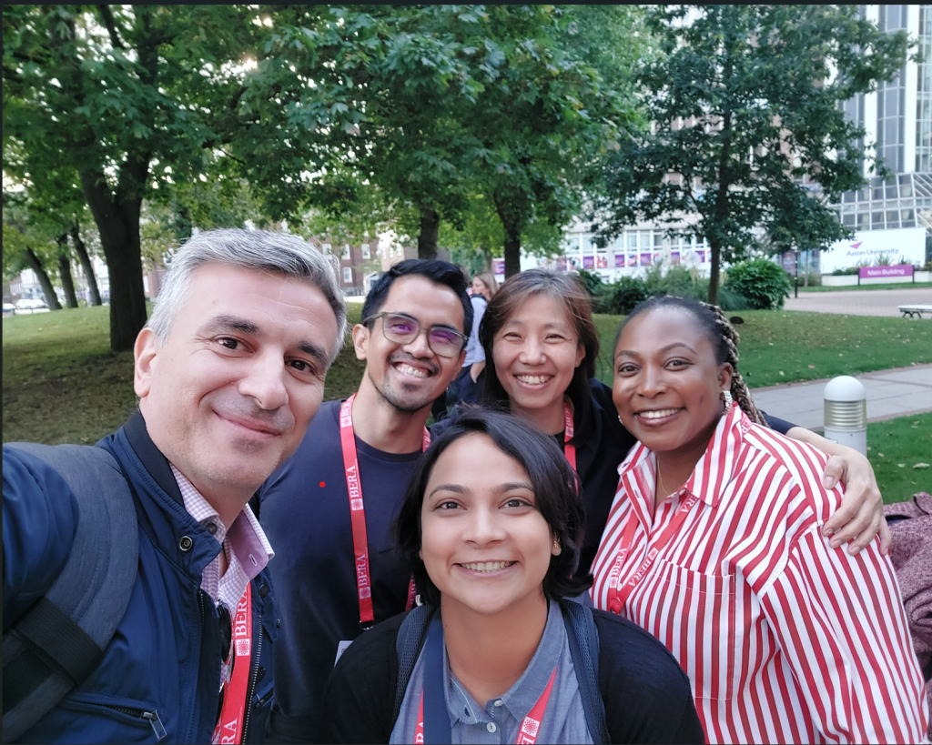 A group photo of 5 people stood outside a building, surrounded by many trees. 