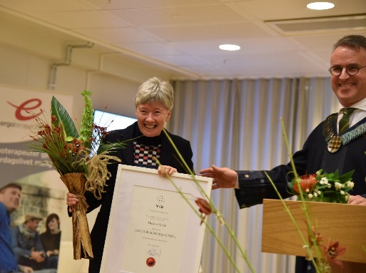 An image of Professor Melanie Nind on a stage, holding a bouquet of flowers and a large, framed certificate