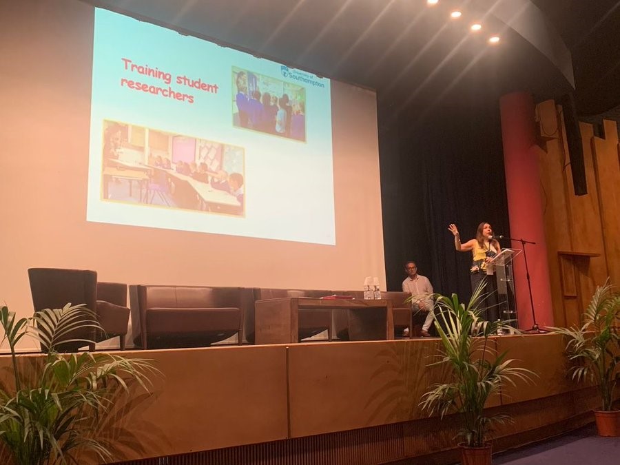 A woman stood at the podium on a stage, pointing at the projector screen with text and images