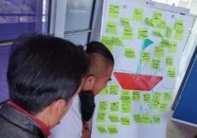 Two people looking at the sticky notes and the boat like structure on a white board