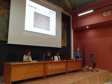 4 people on a panel on the stage, with one person standing and speaking while pointing at the presentation slides projected on the screen behind them.