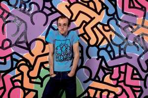 Keith Haring wearing a blue t-shirt standing in front of one of his artworks.