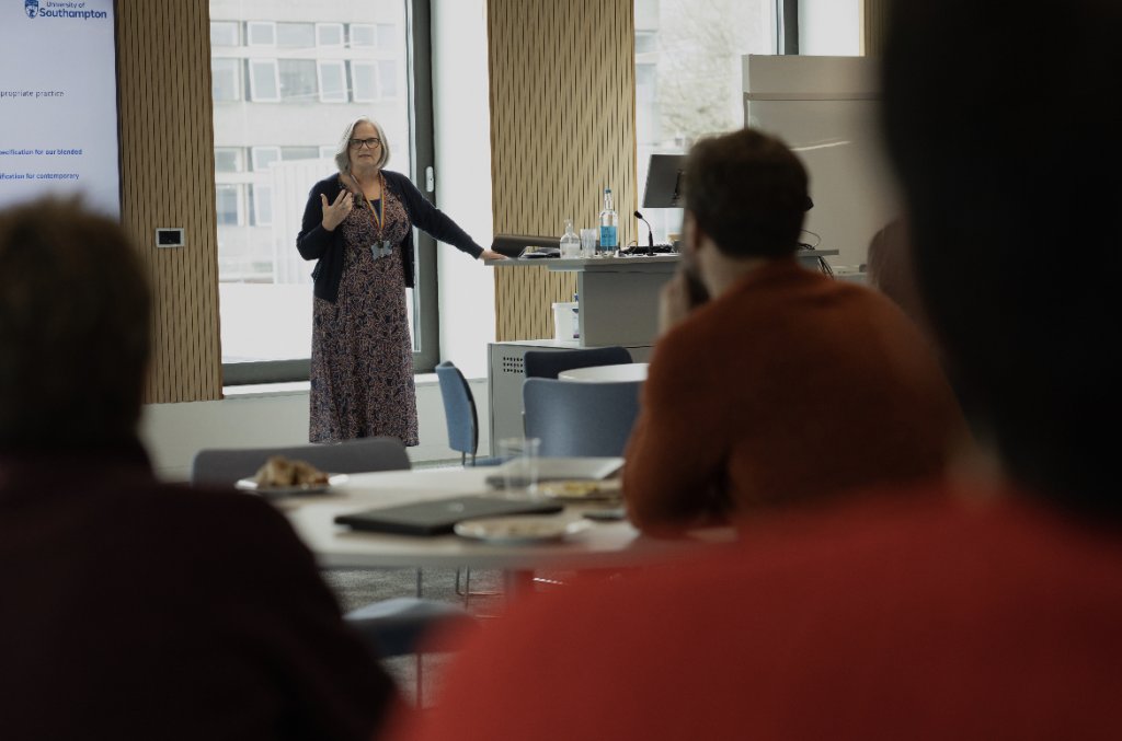 The back view of attendees is blurred out. The camera is focusing on the presenter who stands in front of a tall window.