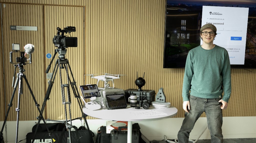 Image contains media devices like the drone, tablet and camera with various  lenses set on a round white table. Left side of table has two camera stands. On the right side of the table  stands a smiling member of the Multimedia team.