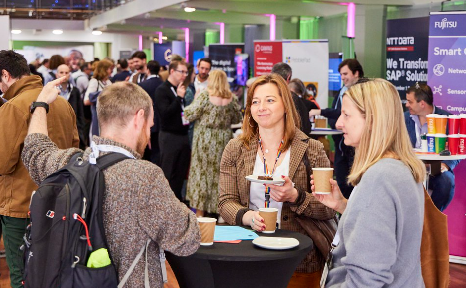 Three delegates chat over lunch in a busy room with educational technology stands at the edges