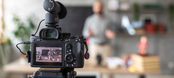 DSLR camera on tripod, recording a video of teacher. He is in the background of the shot, standing behind a table, out of focus.