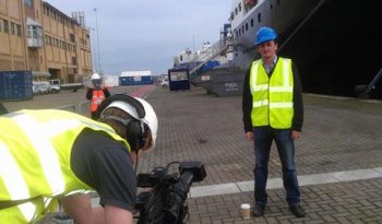 An academic in a hi-vis jacket and hard hat is standing in a port, with a ship in the background. In the foreground, camera operator is filming him.