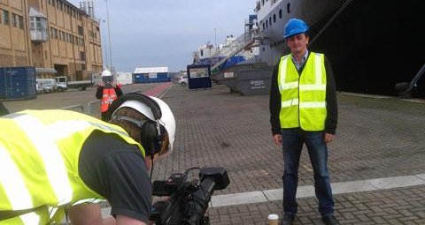 An academic in a hi-vis jacket and hard hat is standing in a port, with a ship in the background. In the foreground, camera operator is filming him.