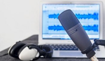 A microphone is in front of a laptop and headphones. The laptop displays audio recording software.