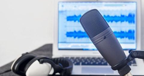 A microphone is in front of a laptop and headphones. The laptop displays audio recording software.