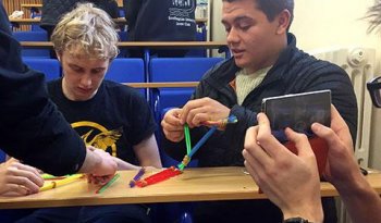 Three students are constructing a model out of straws and elastic bands, while another student shoots this on their phone.