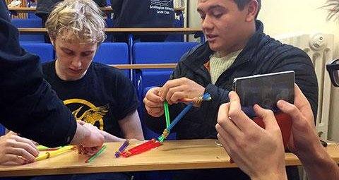 Three students are constructing a model out of straws and elastic bands, while another student shoots this on their phone.