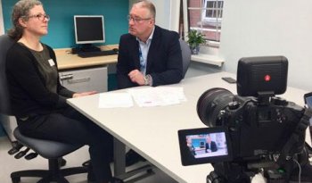 A behind-the-scenes view of shooting an outreach video. A male and female colleague are being filmed having a discussion, seated at a desk with documents in front of them.