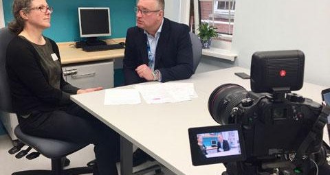 A behind-the-scenes view of shooting an outreach video. A male and female colleague are being filmed having a discussion, seated at a desk with documents in front of them.