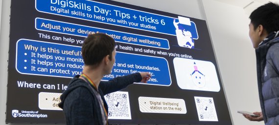 A sideview of James Allen, Senior Learning Designer in the Digital Learning team, talking to a student and pointing at the large digital screen behind him which displays one of the 'Digital tips and tricks' posters for how to adjust your device for digital wellbeing.
