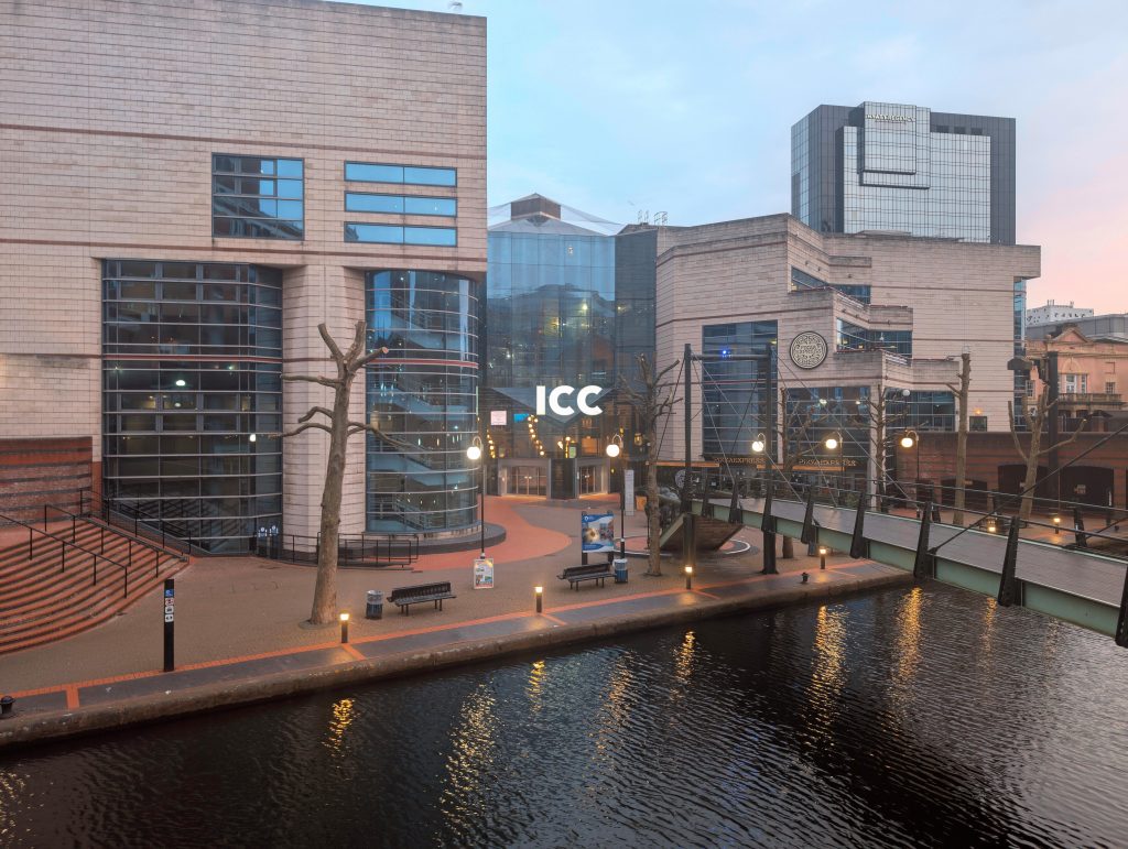 Birmingham's International Convention Centre at dusk. Street lights light the path adjacent to the canal and there is a footbridge to the centre.