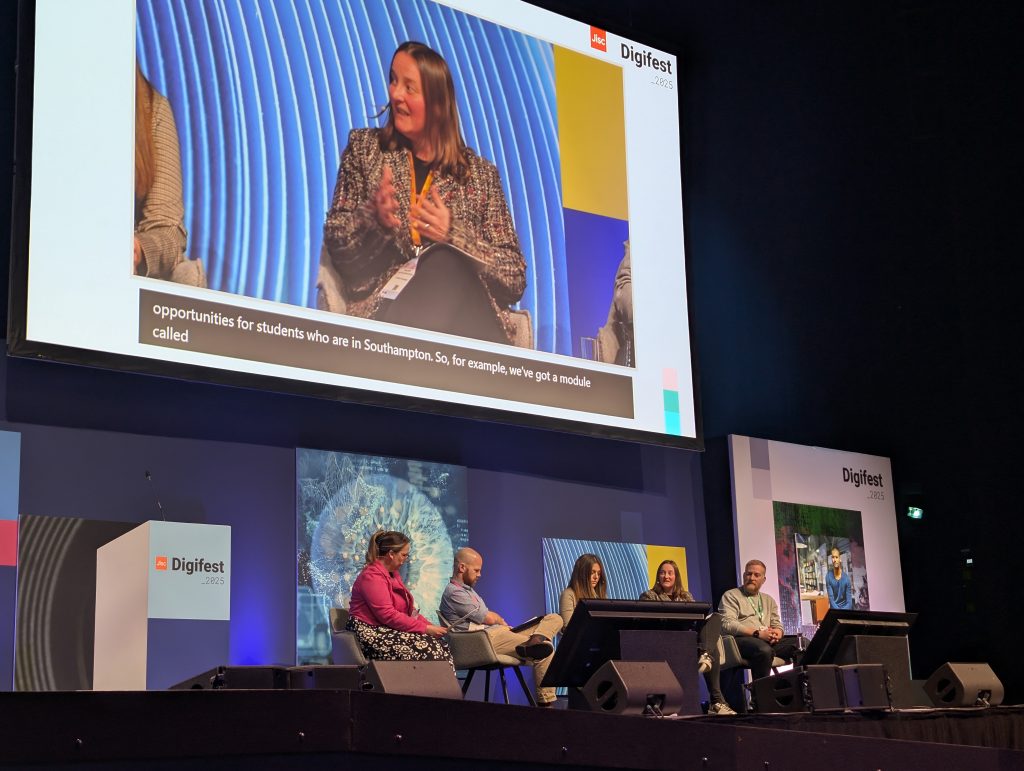 Tamsyn Smith speaking at Digifest, they also appear on a large display above the stage they are sat on.