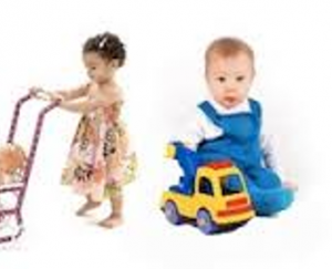 A infant boy is shown to be playing with a toy truck, dressed in blue dungarees. An infant girl is standing and is pushing a pram with a doll inside, dressed in a floral dress.