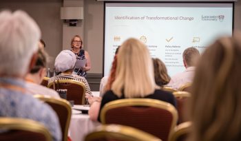Person presenting at a conference in front of a projected slide titled "Identification of Transformational Change," with attendees seated.
