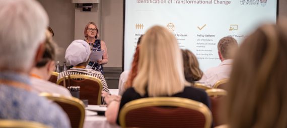 Person presenting at a conference in front of a projected slide titled "Identification of Transformational Change," with attendees seated.