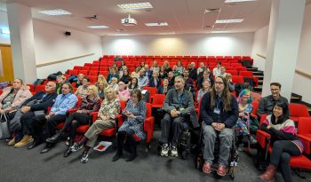 Group photo of approximately 40 presenters and organisers seated in a lecture theatre with red tiered seating. Attendees include wheelchair users in the front row.