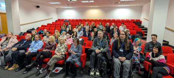 Group photo of approximately 40 presenters and organisers seated in a lecture theatre with red tiered seating. Attendees include wheelchair users in the front row.