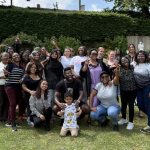 A picture of a group of about 30 happy people (mainly, but not only, women) of different races, religions, and ages in a sunny garden, smiling at the camera and having a good time.
