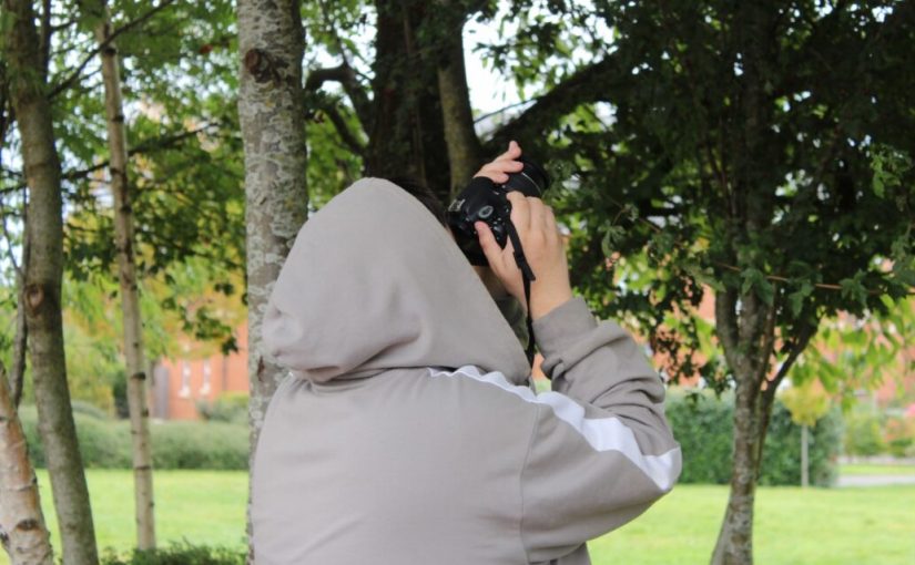 A person with a beige hoodie is pointing their camera towards the trees.