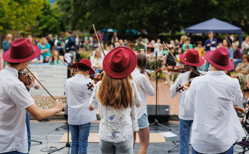 A group of strings players perform outdoors in front of a crowd.