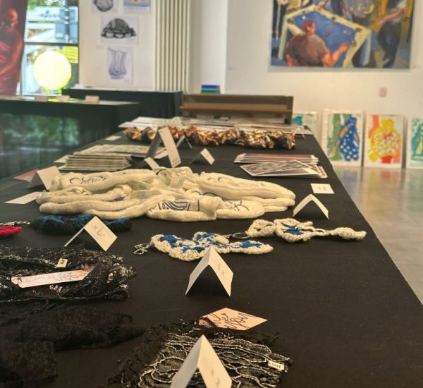 A close-up view of a display table with handmade textile pieces and jewelry, with a large painting and colorful portraits visible in the background.