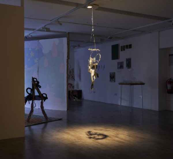 A dimly lit gallery space features a hanging sculpture illuminated by a spotlight, casting a shadow on the floor. In the background, additional artworks and installations are visible against white walls.