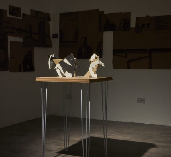A wooden table with thin metal legs displays abstract sculptural pieces made of light-colored materials, illuminated by a spotlight. The background wall features several dark-toned artworks arranged in a collage-like manner.