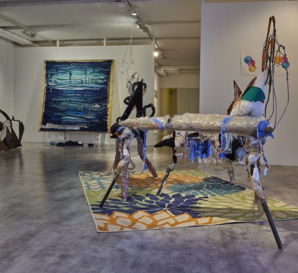 A gallery installation features an abstract sculptural piece made of mixed materials on a colorful floral-patterned rug in the foreground. In the background, a large blue textured artwork hangs on the wall alongside other abstract sculptures and small framed pieces.